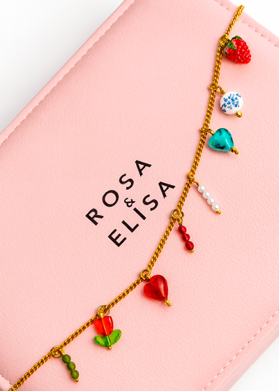 Pulsera dorada con charms y colgantes de colores sobre fondo rosa con el logo Rosa & Elisa, joyería y accesorios en Valencia.